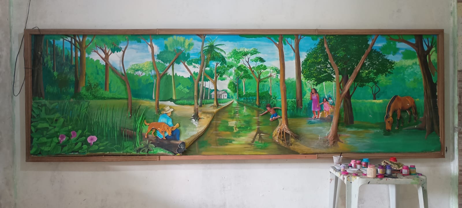 Uma pintura horizontal ampla que mostra um igarapé (pequeno rio) atravessando uma floresta densa. À esquerda, um homem sentado em um tronco caído observa o rio acompanhado de seu cachorro caramelo. À direita, crianças brincam na água rasa e um cavalo marrom bebe água na margem. A luz do sol filtra-se por entre as copas das árvores verdes.
